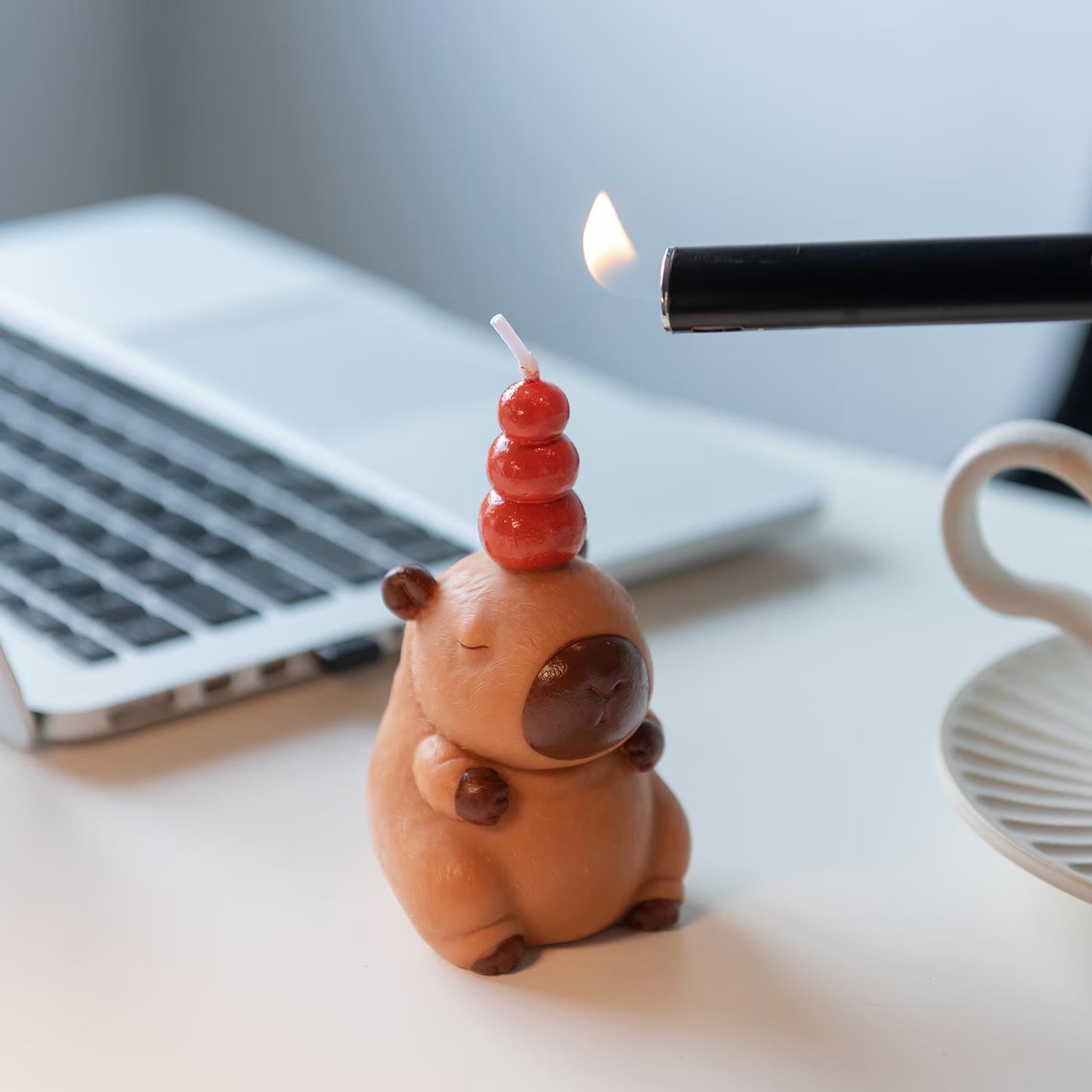 MrsCapybara capybara birthday candle being lit with a lighter on a white desk near a laptop and a coffee cup