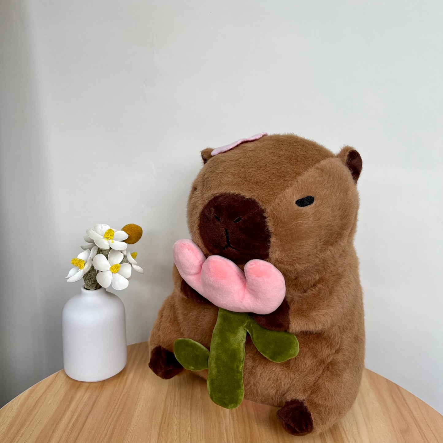 capybara plush with rose sitting on wooden table next to white vase with flowers