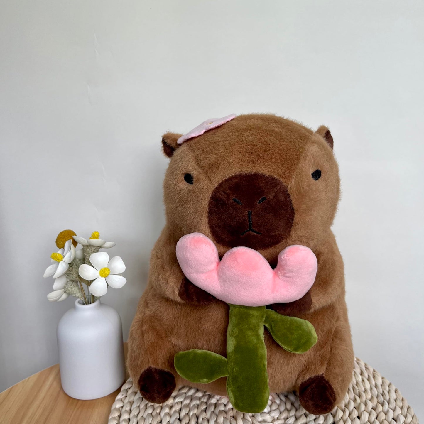 capybara plush with rose holding a pink flower sitting on a woven mat next to a white vase with flowers