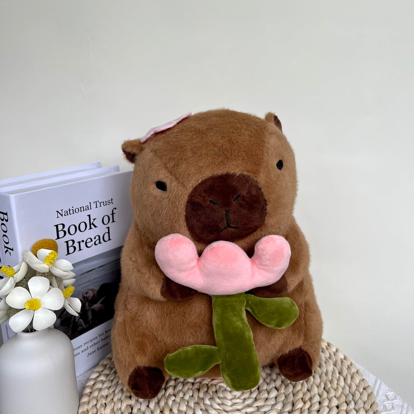 capybara plush with rose holding a pink flower sitting on a woven mat next to books and a vase