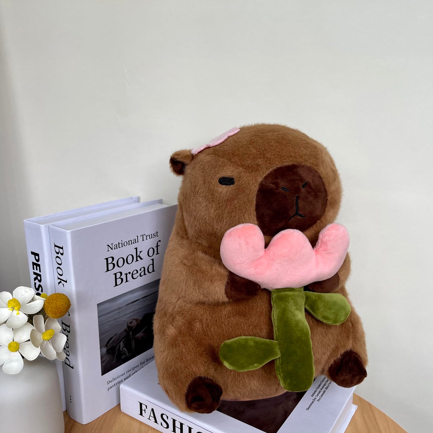capybara plush with rose holding a pink flower sitting on books on a wooden table