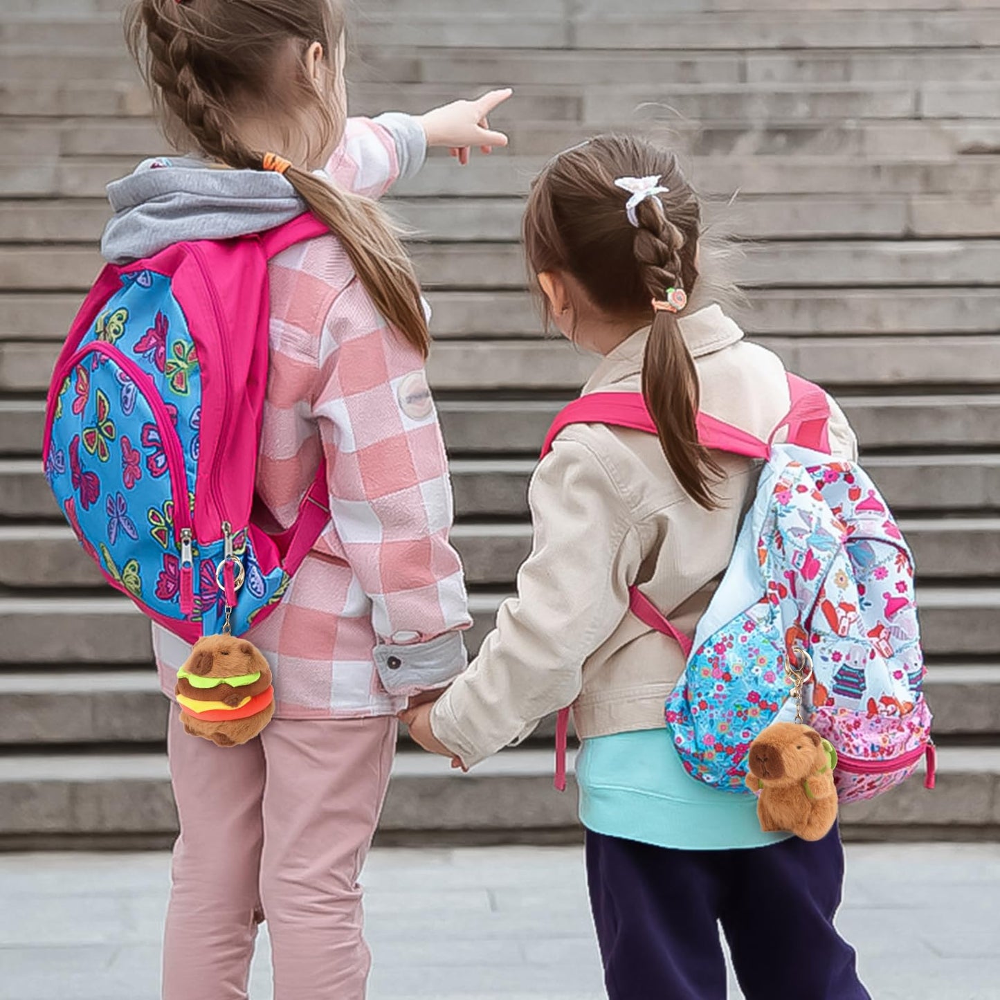 Two children wearing colorful backpacks with plush capybara keychain set attached outdoors near stairs