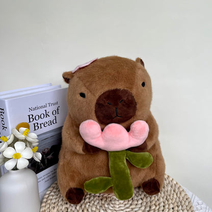 capybara plush with rose holding a pink flower sitting on a woven mat next to books and a vase
