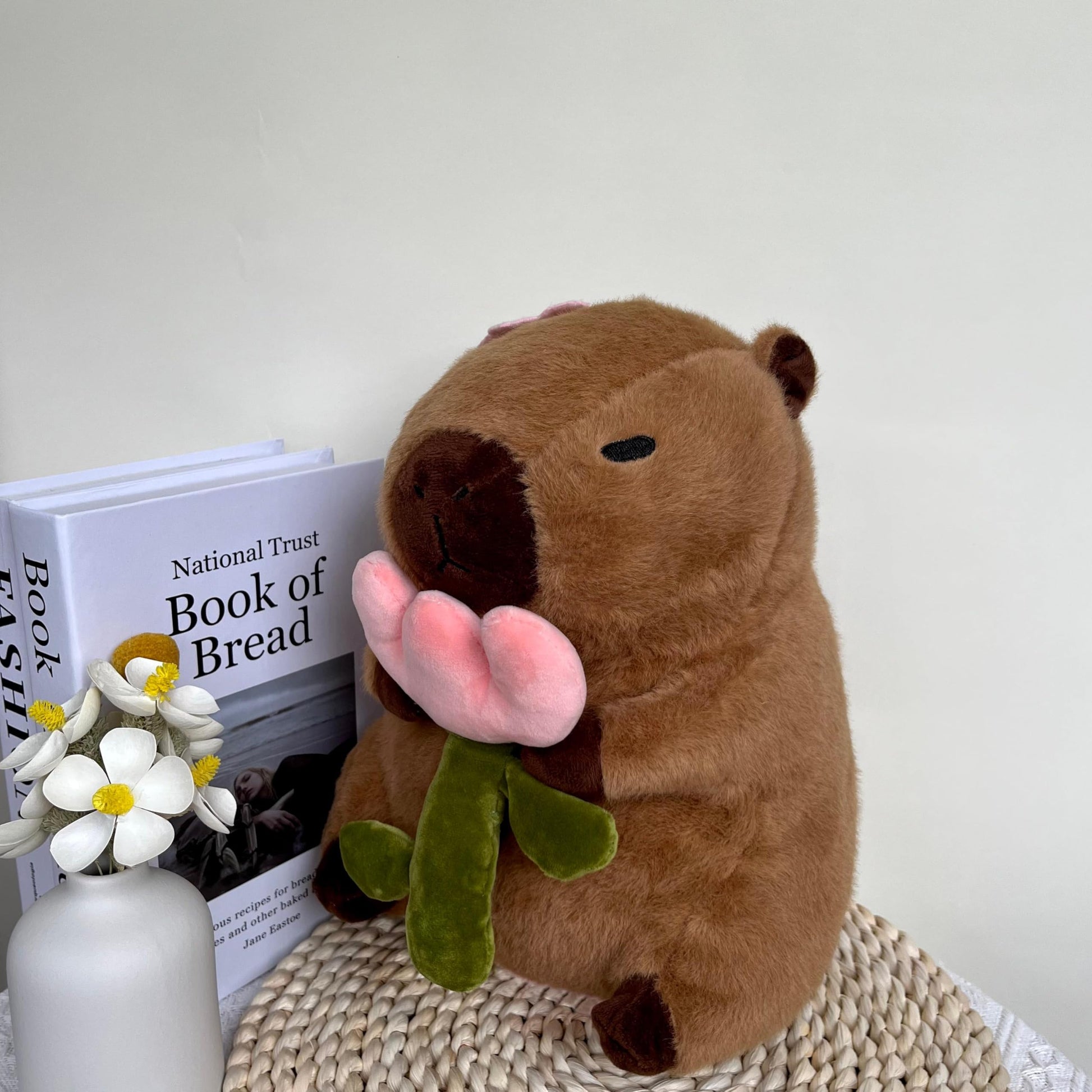 capybara plush with rose holding a soft pink and green flower sitting on a woven surface