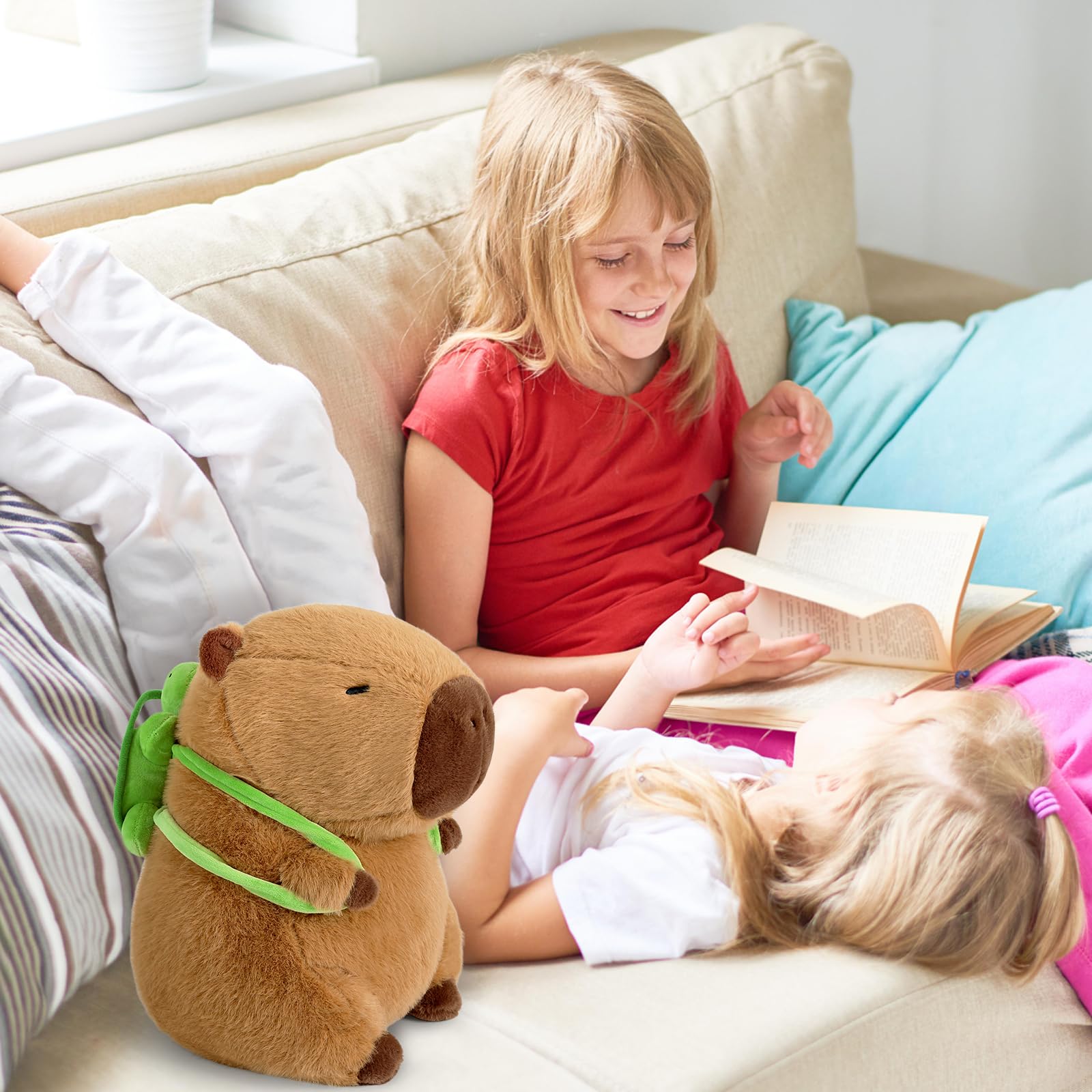 Capybara stuffed animal with turtle backpack sitting on couch near two children reading a book